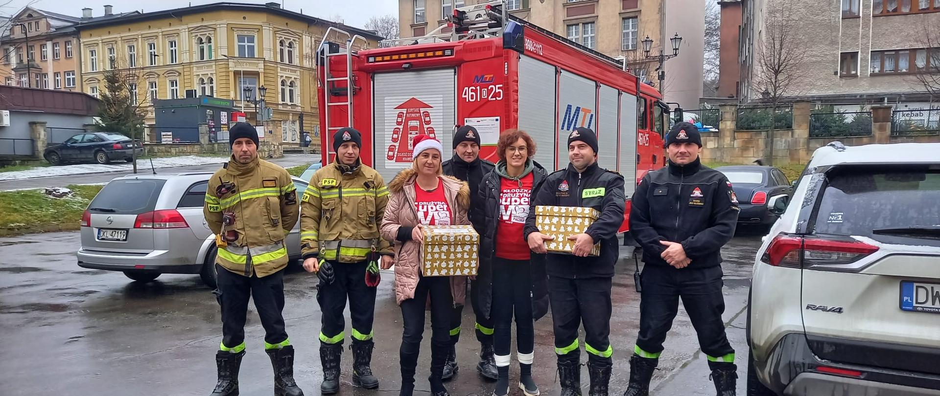 Na parkingu stoi pięciu strażaków i dwie kobiety. Jedna z kobiet oraz jeden ze strażaków trzymają w ręku prezentowe pudełka. 