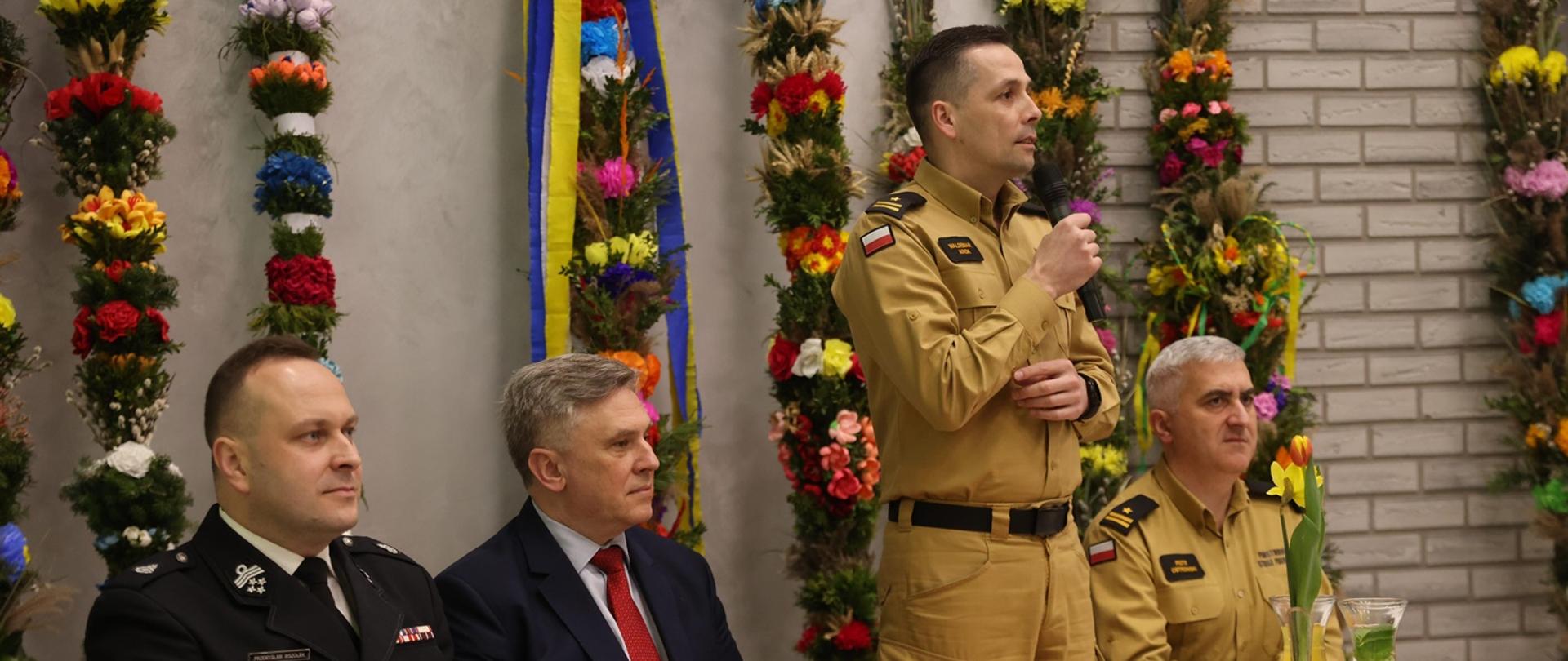 Na zdjęciu widoczny komendant powiatowy Państwowej Straży Pożarnej podczas przemówienia na naradzie operacyjnej dla naczelników OSP i komendantów gminnych ZOSP RP z terenu powiatu gorlickiego. Komendant ubrany w ubranie służbowe koloru pisakowego przemawia obok przy stole siedzą zastępca komendanta powiatowego w Gorlicach ubrani w ubrania służbowe koloru pisakowego, prezes powiatowy Związku Ochotniczych Straży Pożarnych Rzeczypospolitej Polskiej w Gorlicach ubrany w ubranie wyjściowe koloru granatowego, wójt gminy Łużna w garniturze granatowym.