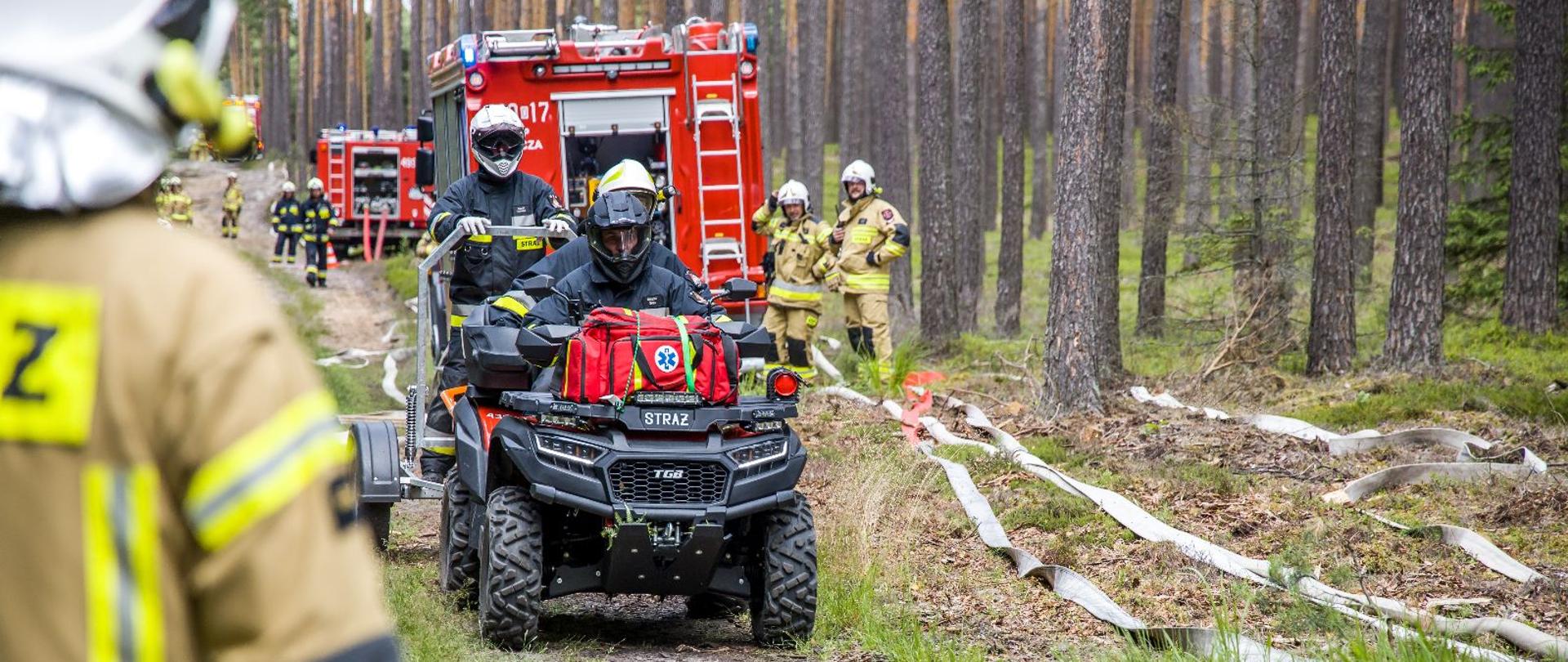 Strażacy w lesie podczas rozwinięcia bojowego. Na pierwszym planie quad transportujący osobę poszkodowaną.