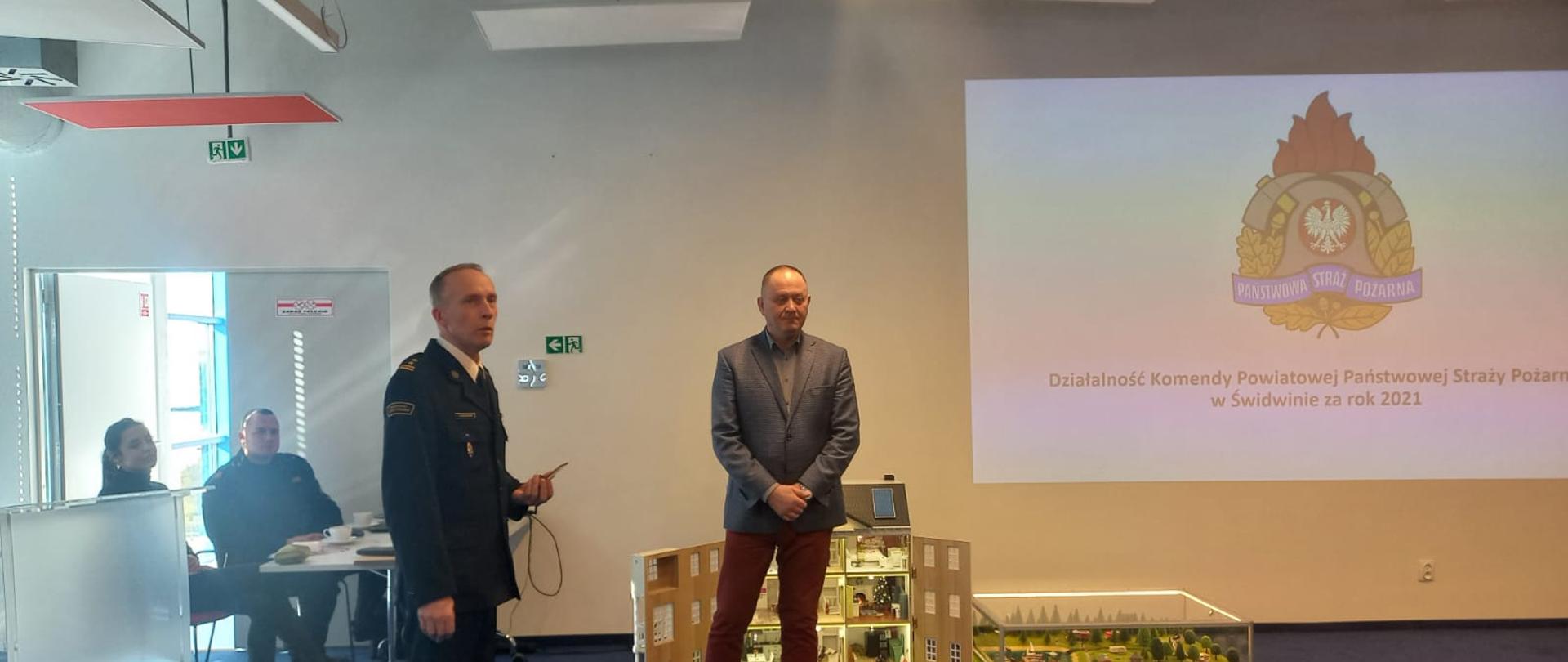 Zdjęcie przedstawia dwóch mężczyzn podczas podsumowanie działalności KP PSP Świdwin za rok 2021 w Centrum Nauki CORDIS, jeden z nich jest w mundurze. Na planszy wyświetlany jest slajd. Obok nich znajduje się makieto domku - symulator pożarów jako stanowisko dydaktyczne w prewencji społecznej. Obok nich znajdują się dwie osoby siedzące przy biurku.