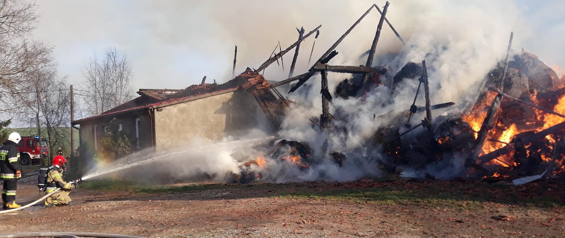 Zdjęcie przedstawia działania gaśnicze w natarciu na palące się pozostałości budynku magazynowego (stodoła ) prowadzone przez dwóch strażaków (jednego z Państwowej Straży Pożarnej, a drugiego z Ochotniczej Straży Pożarnej)
