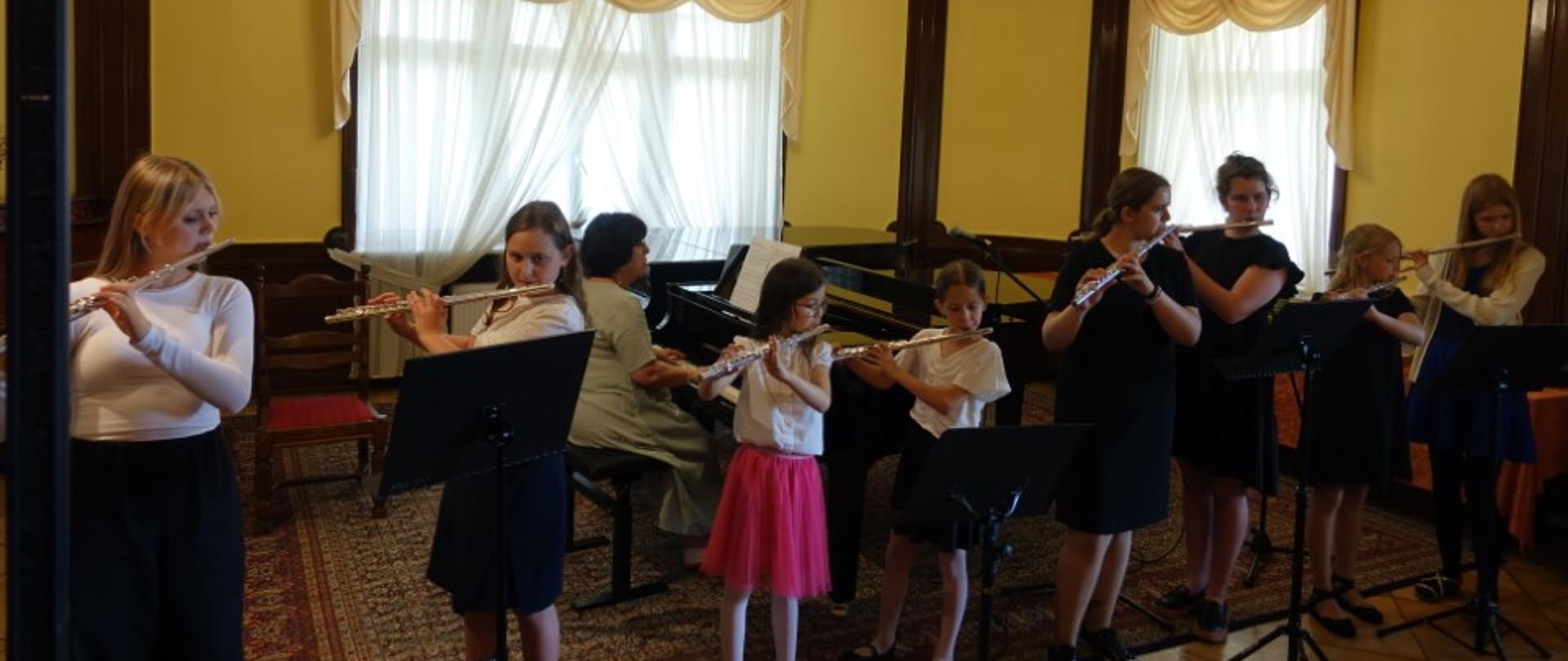 Zdjęcie kolorowe. Grupa dzieci grających na fletach. W tle akompaniator przy fortepianie.