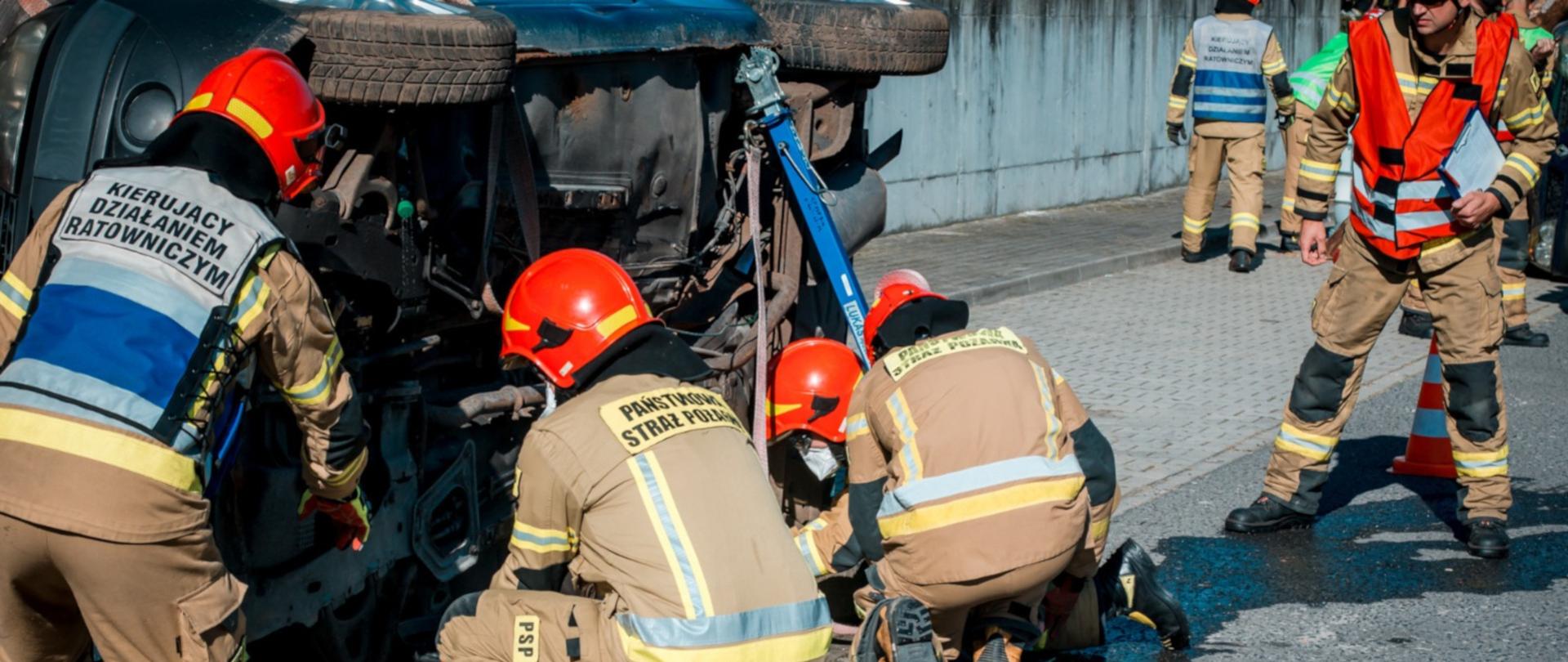 Na zdjęciu czterech strażaków w ubraniach specjalnych dokonuje stabilizacji pojazdu przewróconego na bok. Obok stoi strażak w ubraniu specjalnym - sędzia