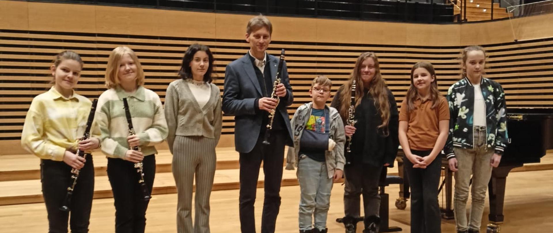 Ośmiu uczestników warsztatów i prowadzący stoją na scenie sali koncertowej PSM, trzy uczennice i prowadzący trzymają klarnety.