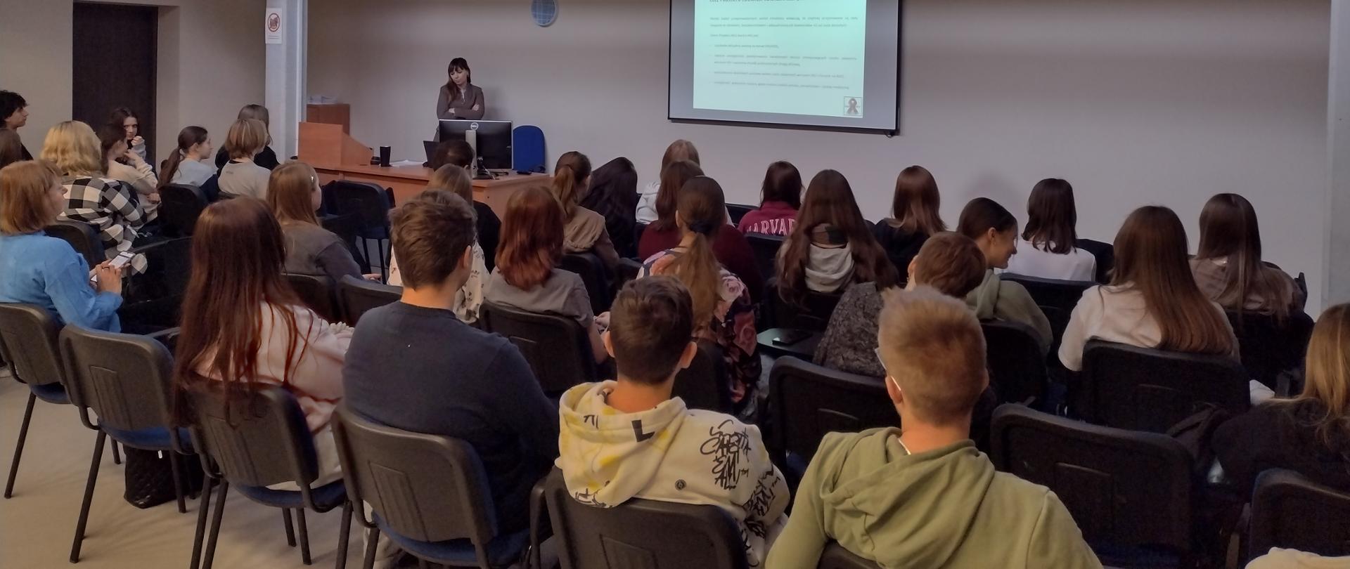 Na pierwszym planie widoczny jest tłum osób, którzy znajdują się w sali wykładowej. W tle widać kobietę, która przemawia.