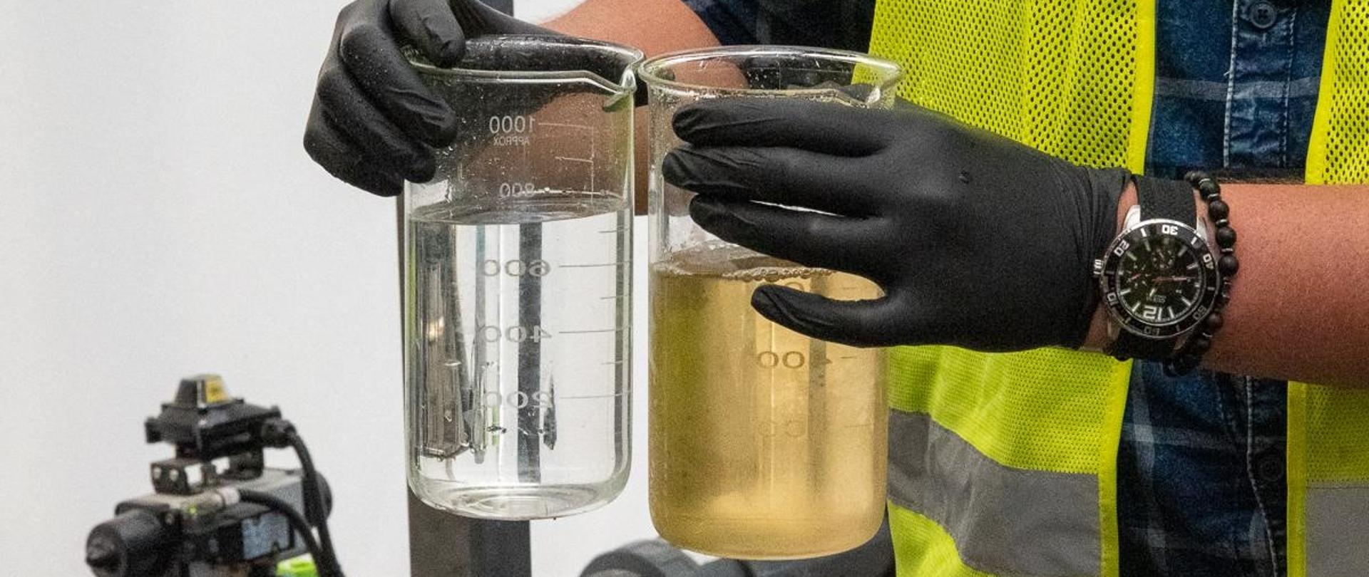 Mężczyzna w żółtej kamizelce trzyma dwie próbki wody - mętną i czystą
