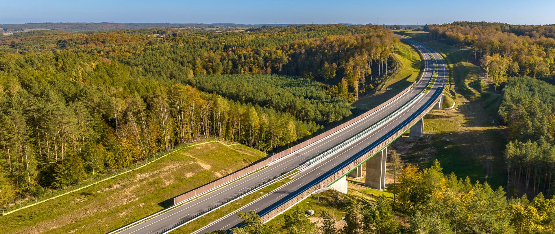 Zdjęcie przedstawia drogę ekspresową S6 na odcinku Luzino - Szemud. Widok z lotu ptaka ukazuje nowoczesną, wielopasmową drogę przecinającą malowniczy krajobraz pełen zieleni i lasów. Droga biegnie przez most, który wznosi się nad doliną, co dodaje zdjęciu dynamiki i ukazuje infrastrukturę wkomponowaną w naturalne otoczenie. Na horyzoncie widoczny jest rozległy obszar leśny, a niebo jest bezchmurne i jasnoniebieskie, co podkreśla piękno dnia. 