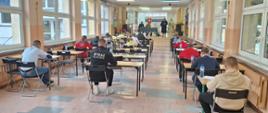 Zdjęcie przedstawia uczestników turnieju wiedzy pożarniczej którzy siedzą na sali podczas pisania testu. Komisja siedzi przy stoliku na podwyższeniu. 