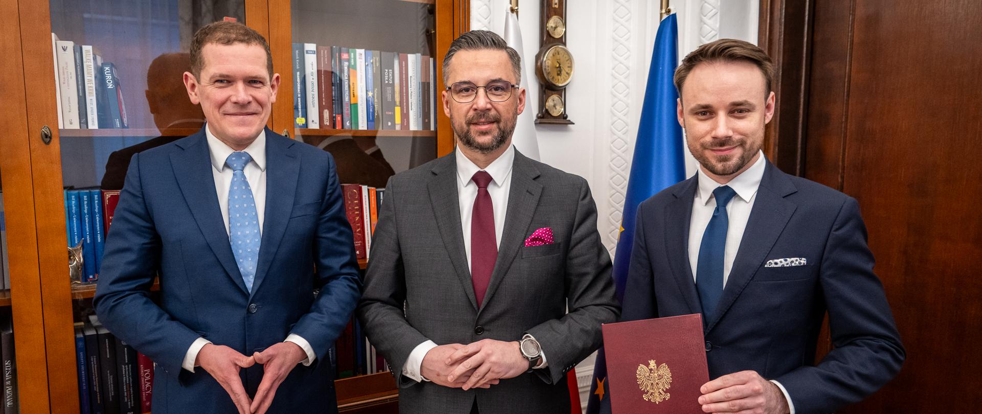 W gabinecie stoi trzech mężczyzn w garniturach, pośrodku minister Kulasek, z prawej Damian Syjczak trzyma w ręku czerwoną teczkę, za nimi przeszklona szafa z książkami.