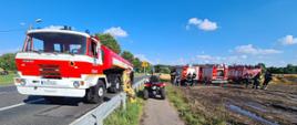 Na zdjęciu widać Drogę Krajową nr 12. W centralnym punkcie samochód cysterna. Posiada kolor czerwono-biały. Na dachu sygnalizacja ostrzegawcza. Po prawej, na polu samochody gaśnicze oraz quad również w kolorze czerwonym. Obok quada strażak ubrany w umundurowanie specjalne w kolorze pisakowym. Niebo lekko zachmurzone. 