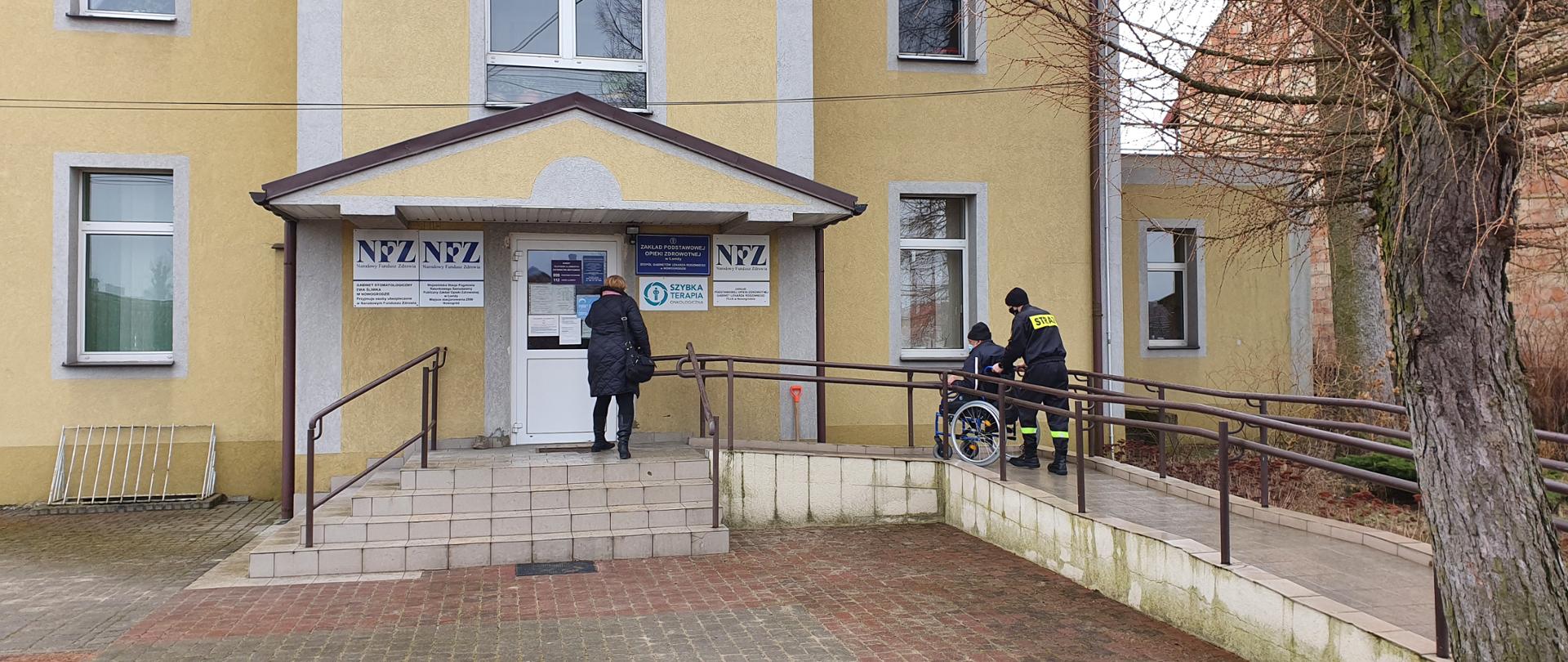 Na tle budynku przychodni lekarskiej obecne są trzy osoby. Z prawej strony widoczni na podjeździe dla osób niepełnosprawnych są dwaj mężczyźni: jeden będący na wózku inwalidzkim pchanym przez strażaka. Drzwi budynku otwiera im kobieta. 