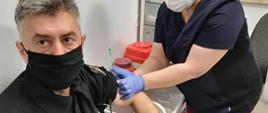 Zdjęcie przedstawia strażaka poddającego się szczepieniu przez naczelną pielęgniarką Szpitala w Opatowie. Strażak i pielęgniarka w maseczkach ochronnych oraz w rękawiczkach nitrylowych.