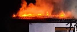 Zdjęcie przedstawia pożar budynku mieszkalnego jednorodzinnego. Płomienie wydostają się z poddasza budynku. 