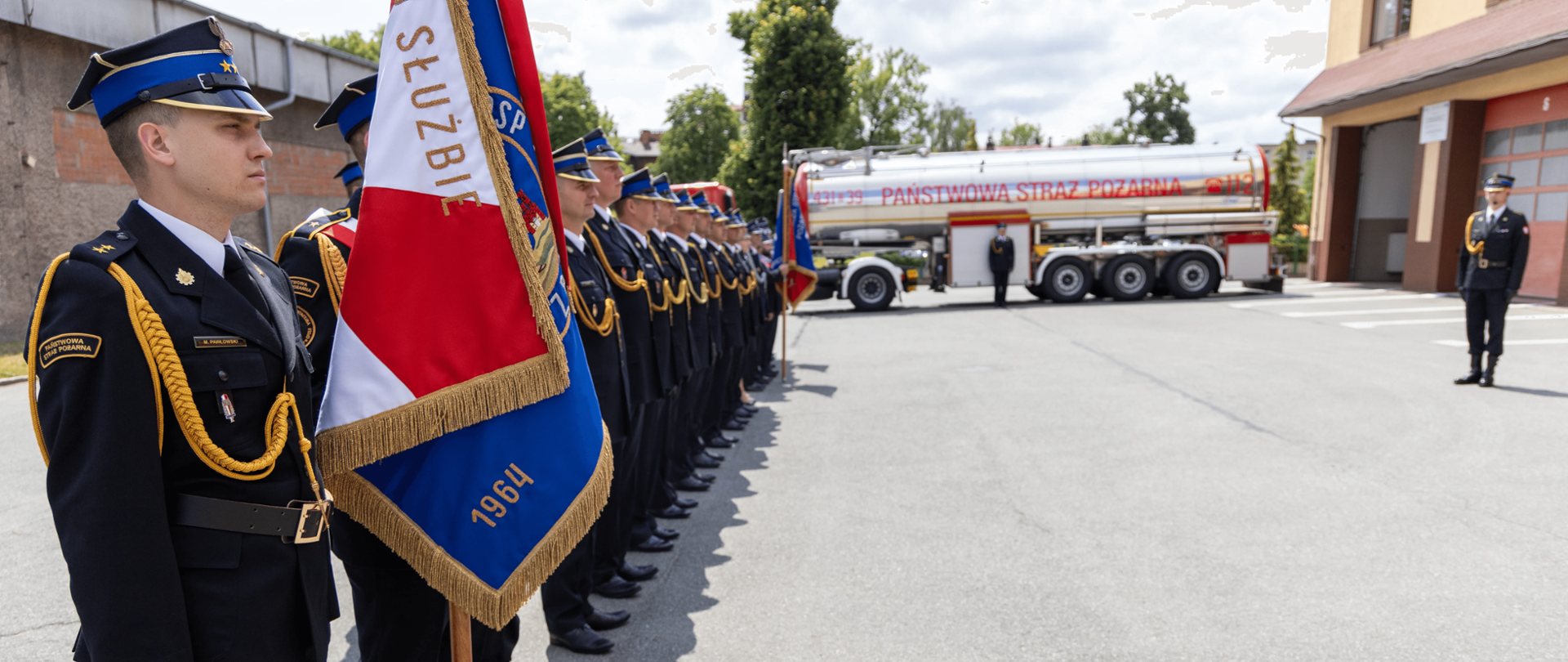 Na zdjęciu znajduje się grupa strażaków w mundurach galowych stojących w szeregu podczas uroczystości. Na pierwszym planie po lewej stronie widać strażaka trzymającego sztandar z czerwono-niebieskim płatem, na którym widnieją złote napisy oraz rok 1964. W tle znajduje się duży wóz strażacki z napisem „PAŃSTWOWA STRAŻ POŻARNA”. Uroczystość odbywa się na placu przed budynkiem strażackim, a w tle widać drzewa i fragment budynku z otwartymi bramami garażowymi. Na zdjęciu panuje pogodna, słoneczna aura.