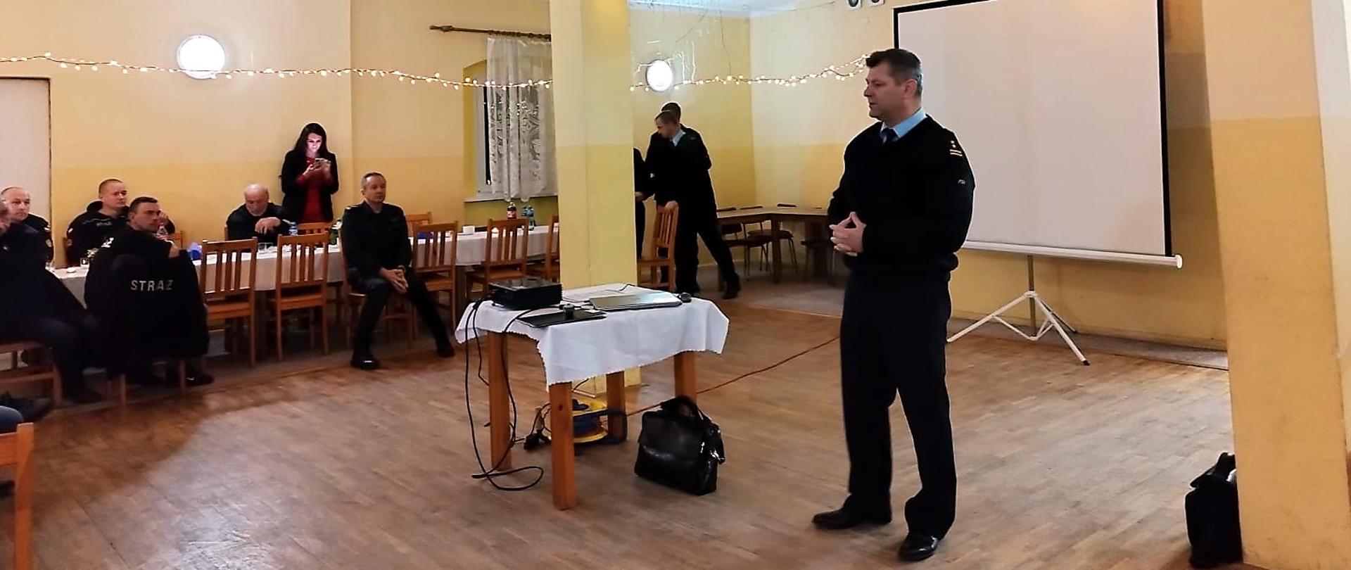 Obraz przedstawia Komendanta Powiatowego PSP w Jaworze brygadiera Waldemara Sarlej przemawiającego podczas odprawy szkoleniowej Prezesów i Naczelników OSP. Osoby w pomieszczeniu świetlicy wiejskiej w Marcinowicach.