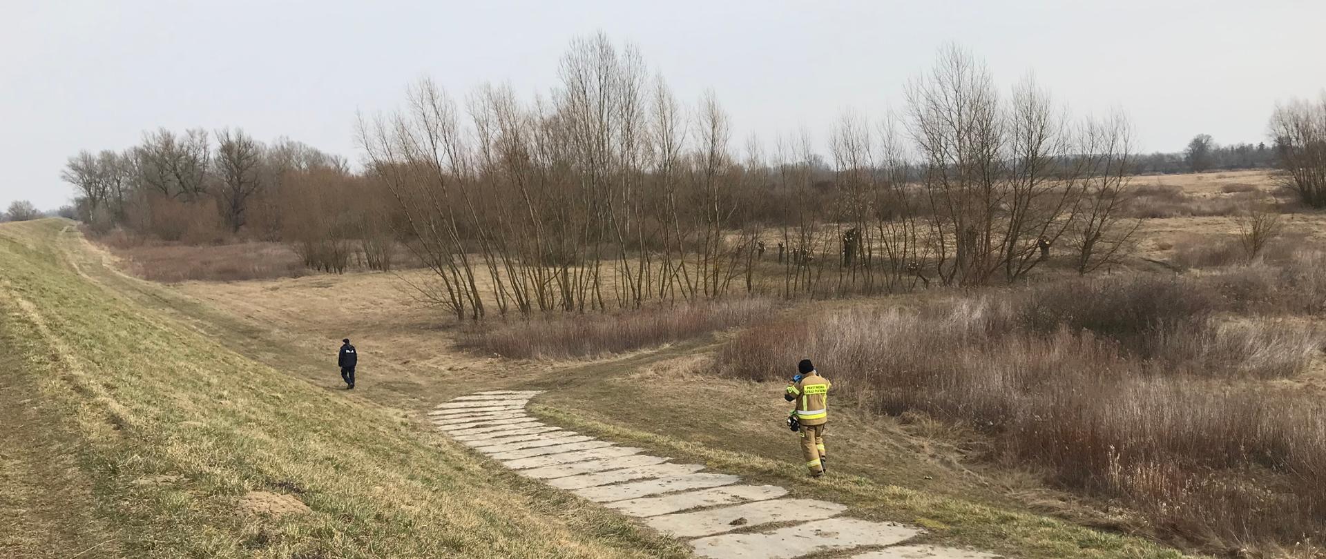 Zdjęcie przedstawia funkcjonariusza Policji oraz strażaka podczas poszukiwań wzdłuż wału przeciwpowodziowego, widoczna jest droga z płyt betonowych oraz tereny nadwiślańskie