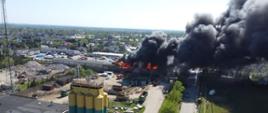 Zdjęcie przedstawia widok z lotu ptaka na przemysłową okolicę. Płomienie obejmują część hali. Nad pożarem unosi się słup czarnego gęstego dymu i rozprzestrzenia zgodnie z kierunkiem wiatru.