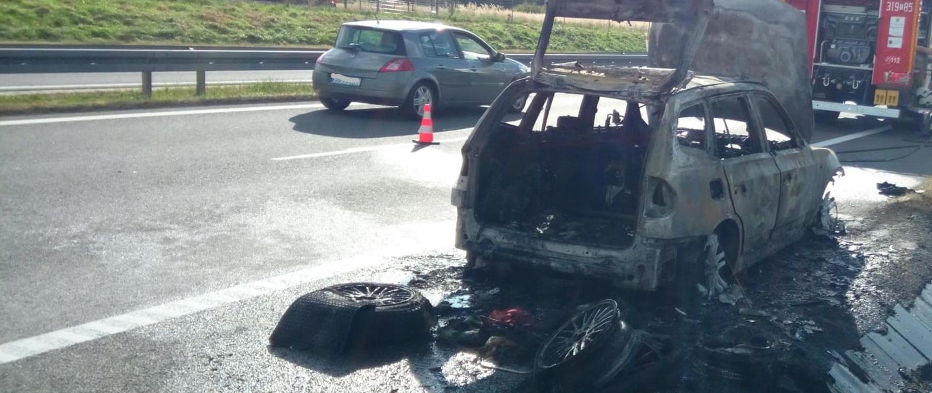 Na zdjęciu, po prawej stronie stoi spalony samochód osobowy, a przed nim samochód pożarniczy. Sąsiednim pasem jedzie przypadkowe auto.
