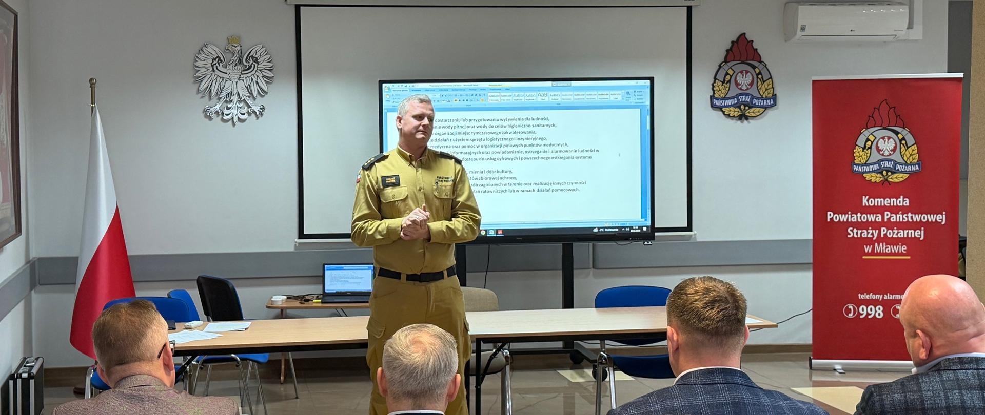 Oficer Państwowej Straży Pożarnej przemawia do zebranych z przodu sali konferencyjnej. Za nim znajduje się ekran z wyświetloną prezentacją tekstową, a po prawej stronie banner Komendy Powiatowej PSP w Mławie. Uczestnicy, siedzący tyłem do obiektywu, notują i przysłuchują się wystąpieniu.