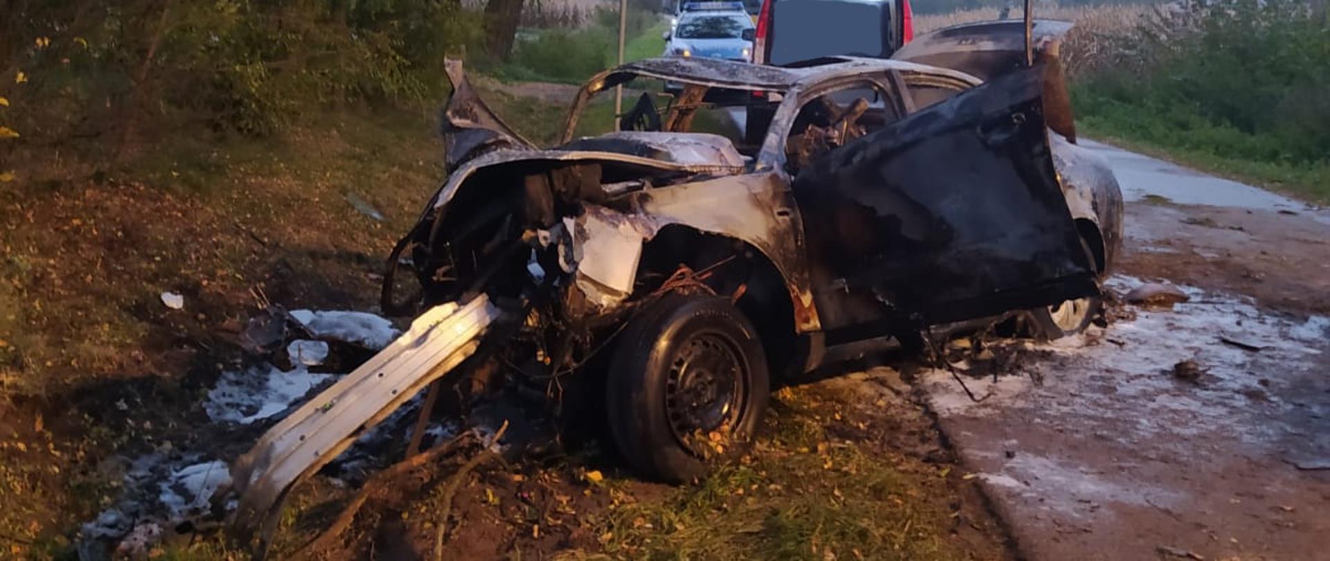 Uszkodzony samochód osobowy po pożarze stoi na poboczu. Za nim stoją samochody służb.