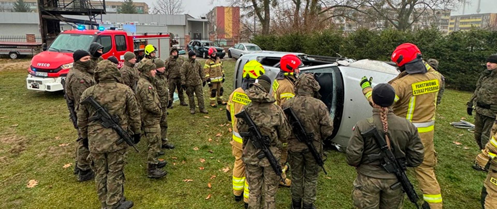 Żołnierze stoją w trakcie szkolenia a w tle przewrócone na bok samochody osobowe, przy których pracują strażacy