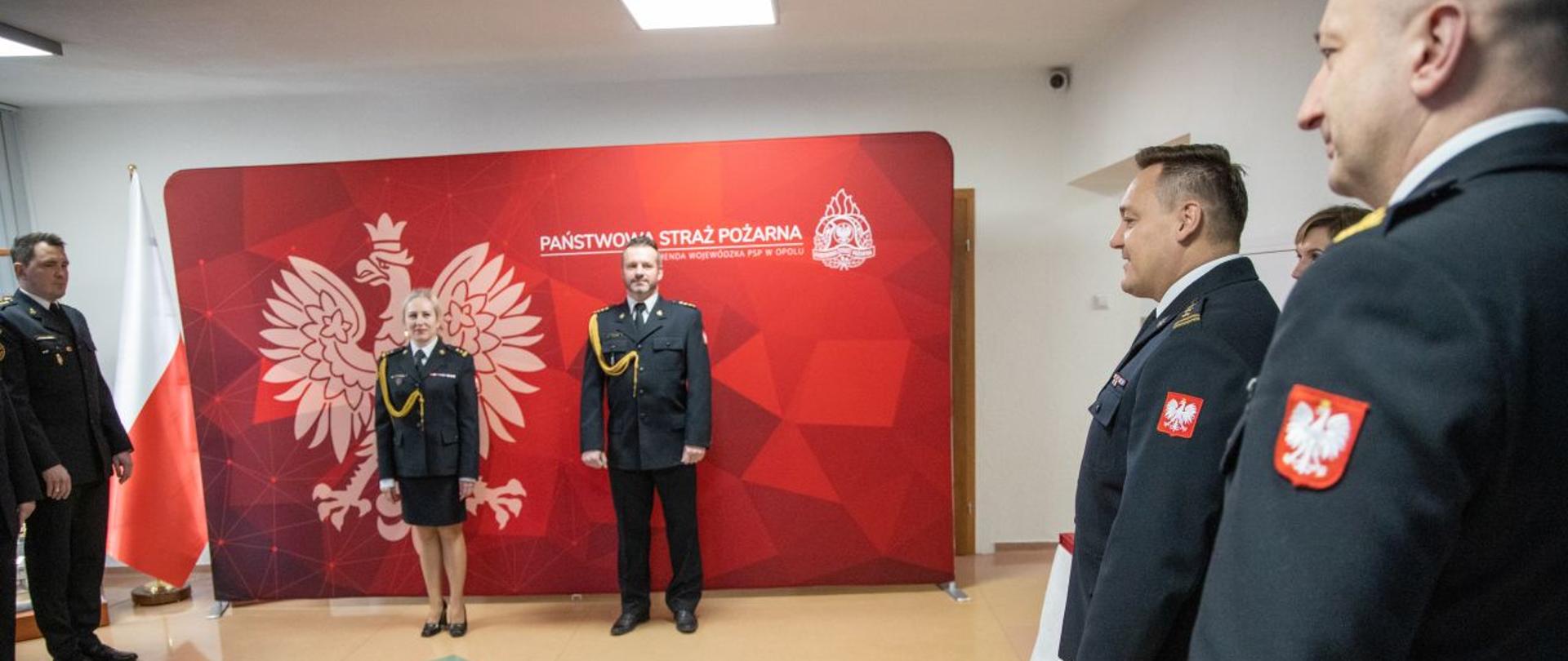 W Komendzie Wojewódzkiej Państwowej Straży Pożarnej odbyła się uroczysta zbiórka strażaków żegnających dwóch funkcjonariuszy przechodzących na zaopatrzenie emerytalne: st. kpt. Romana Felusiaka naczelnika wydziału planowania operacyjnego KW PSP w Opolu oraz
bryg. Dorotę Granacką dyżurną operacyjną Stanowiska Kierowania Opolskiego Komendanta Wojewódzkiego PSP. Strażacy w umundurowaniu galowym na tle ścianki koloru czerwonego z godłem RP i napisem białym Państwowa Straż Pożarna i Komenda Wojewódzka PSP w Opolu oraz logiem PSP.
Po prawie Opolski Komendant Wojewódzki PSP wraz z zastępcą.