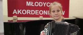 Zdjęcie przedstawia dziewczynkę z akordeonem na tle plakatu II Ogólnopolskich spotkań młodych akordeonistów