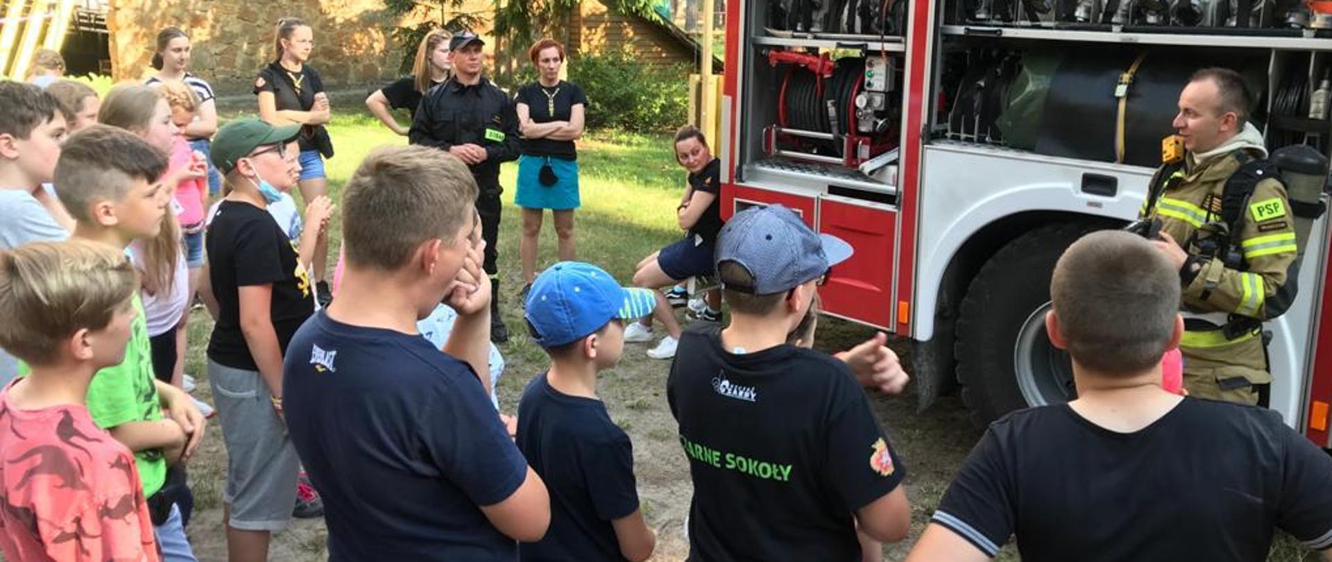 Teren obozu harcerskiego, strażak ratownik prezentuje wyposażenie samochodu ratowniczo-gaśniczego, harcerze zebrani wokół pojazdu, strażak ubrany u ubranie specjalne