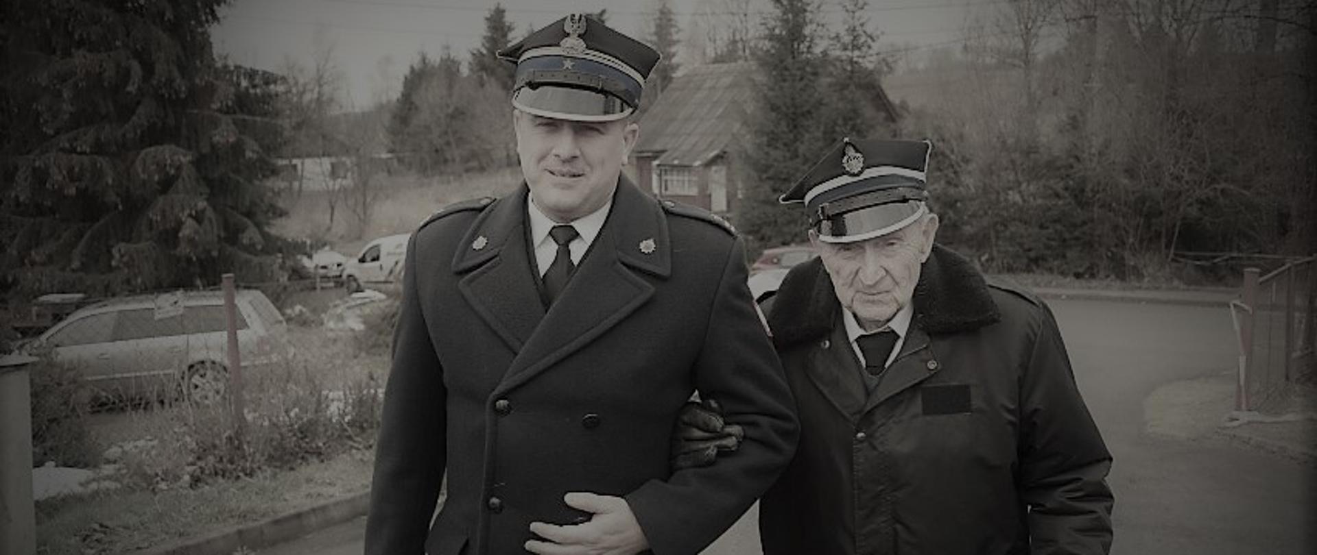 Zdjęcie przedstawia Komendanta Powiatowego PSP w Ustrzykach Dolnych mł. bryg. Emila Szmyda wraz z dh. Franciszkiem Flakiem którego prowadzi pod rękę. Oboje ubrani są w mundury galowe. Druh Franciszek Flak ma ubraną kurtkę i rękawiczki skórzane. W oddali widać samochód osobowy i dom. 