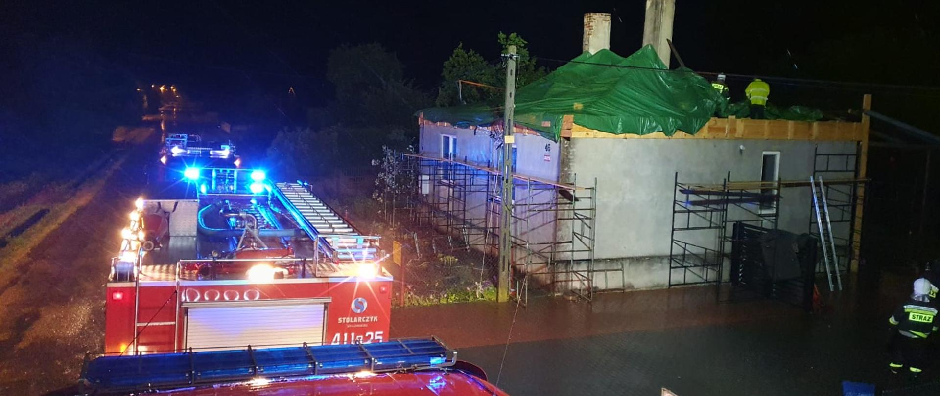 Zdjęcie zrobione w porze nocnej podczas opadów deszczu z dachu samochodu ratowniczo-gaśniczego. Po prawej stronie znajduje się budynek mieszkalny z uszkodzonym dachem. Strażacy pracujący na stropie budynku zabezpieczają go folią przed ponownym zalaniem. Na drodze widnieją zaparkowane pozostałe pojazdy strażackie. 