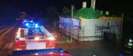 Zdjęcie zrobione w porze nocnej podczas opadów deszczu z dachu samochodu ratowniczo-gaśniczego. Po prawej stronie znajduje się budynek mieszkalny z uszkodzonym dachem. Strażacy pracujący na stropie budynku zabezpieczają go folią przed ponownym zalaniem. Na drodze widnieją zaparkowane pozostałe pojazdy strażackie. 