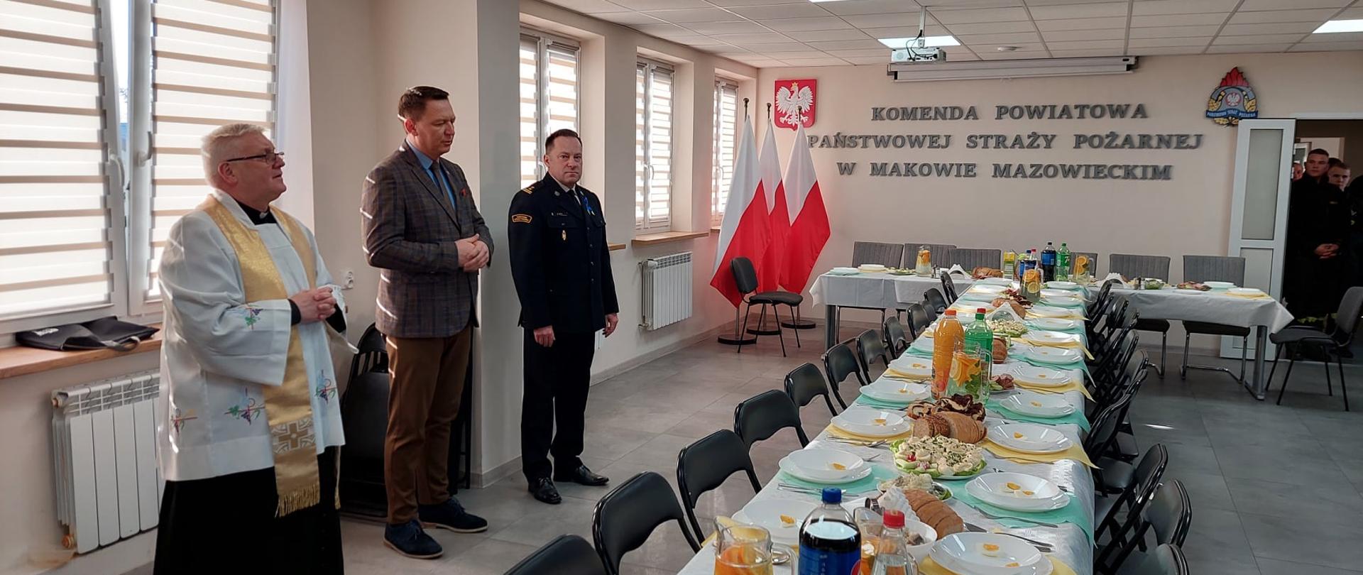 Komendant Powiatowy PSP, Wicestarosta oraz ksiądz kapelan stoją przy wigilijnym stole. W tle napis "Komenda Powiatowa Państwowej Straży Pożarnej w Makowie Mazowieckim", godło Polski oraz logo PSP