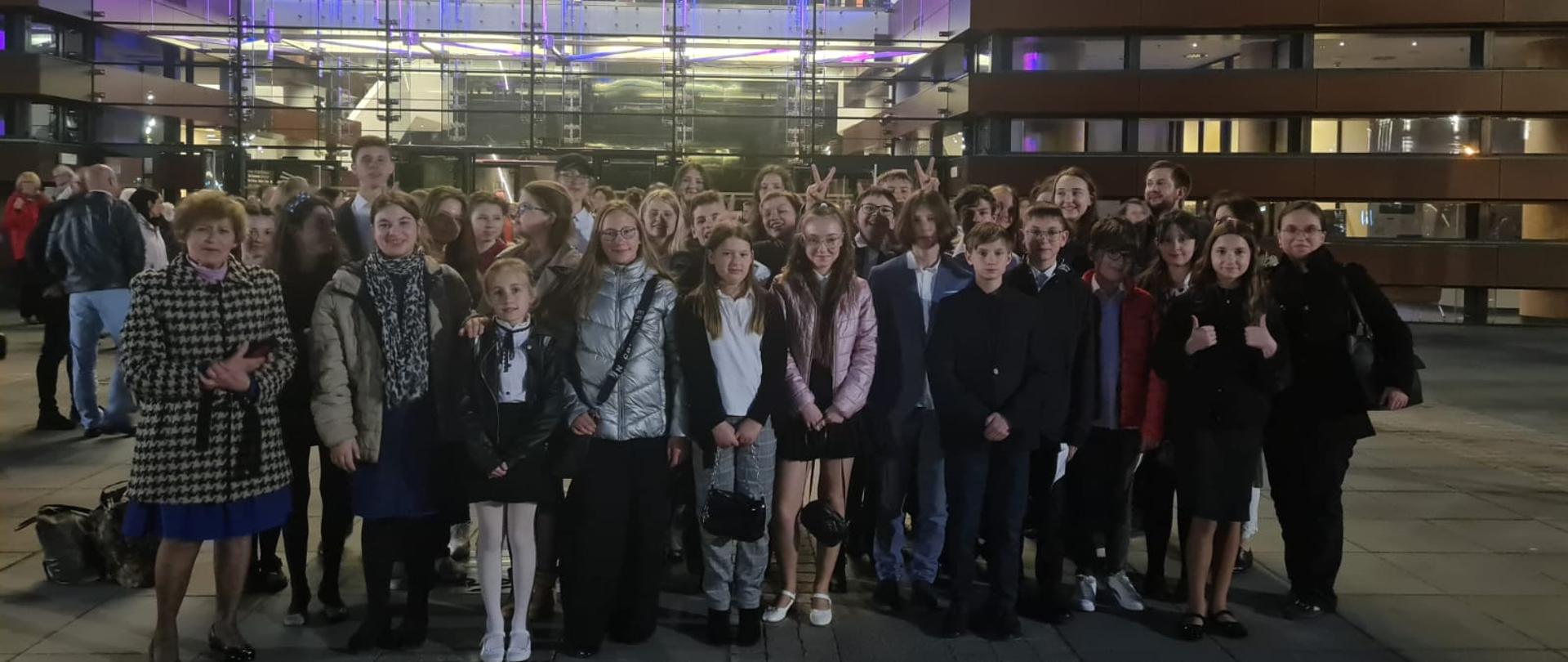 Zdjęcie na zewnątrz przed frontową przeszkloną ścianą Narodowego Forum Muzyki we Wrocławiu. Grupa uczniów Państwowej Szkoły Muzycznej I stopnia w Oławie wraz z nauczycielami i opiekunami.