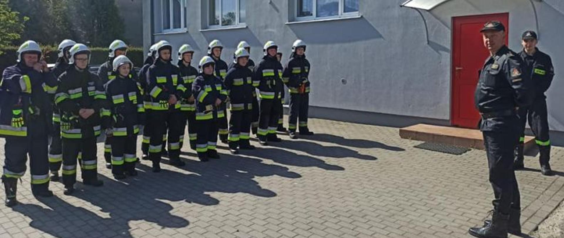 Zbiórka strażaków przed rozpoczęciem szkolenia. Strażacy stoją w ciemnych mundurach i białych hełmach. Przed nimi stoi Dowódca Jednostki, który przekazuje im informacje odnośnie szkolenia.