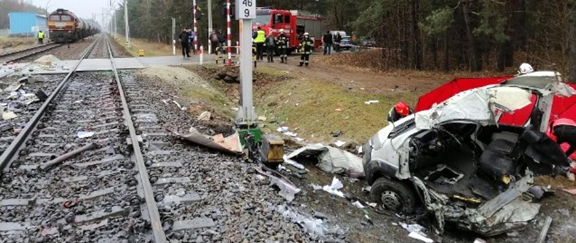 Zdjęcie zrobione przy torach kolejowych. W prawym dolnym rogu zdjęcia znajduje się przód rozbitego samochodu dostawczego. W tle widać samochód strażacki oraz strażaków i policjantów. W lewym górnym rogu widać pociąg stojący na torach. 