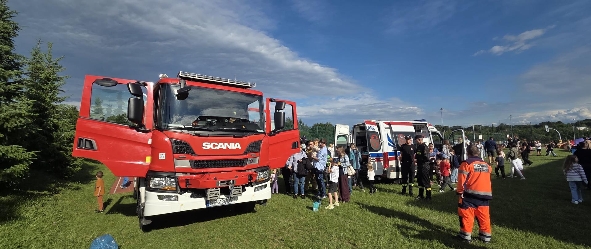 Szeroki kadr przedstawia plenerowy pokaz służb ratunkowych na trawiastym terenie, gdzie po lewej stronie stoi duży, czerwony wóz strażacki Scania z otwartymi drzwiami, a w głębi biało-ordynowany ambulans. Wokół pojazdów gromadzi się tłum dorosłych i dzieci, a na pierwszym planie po prawej stronie tyłem stoi ratownik medyczny w pomarańczowo-czarnym kombinezonie. Nad całą sceną rozciąga się dynamiczne, częściowo zachmurzone niebo w słoneczny dzień.
