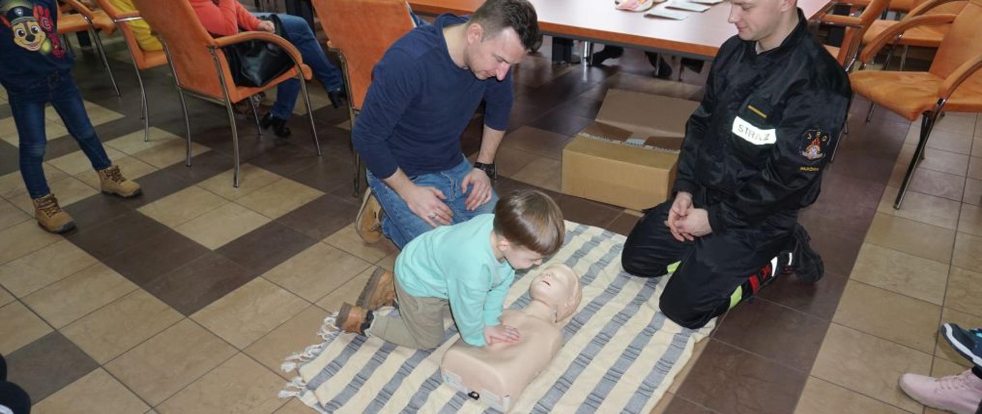 Na podłodze na kocu leży manekin do ćwiczeń resuscytacji. Przy manekinie chłopczyk i dwoje dorosłych. Jeden ubrany w strażacki mundur.