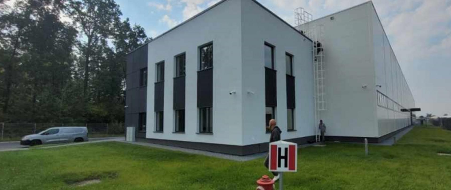 Dwupiętrowy budynek z drabiną, osobami w pobliżu i zaparkowanym vanem; na trawniku hydrant i znak „H”.