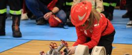 II Wojewódzkie Halowe Młodzieżowe Zawody Sportowo-Pożarnicze - dziewczynka w czerwonym plastikowym hełmie podłącza prądownicę do węża strażackiego siedząc na podłodze