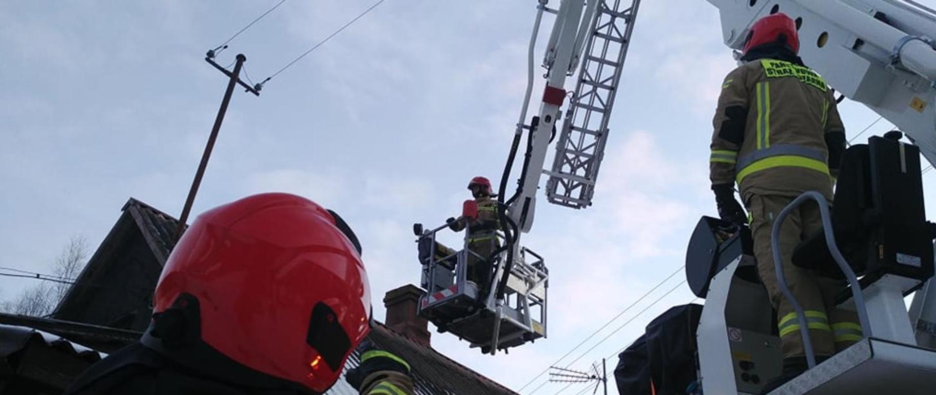 Zdjęcie przedstawia fragment akcji ratowniczej z perspektywy nisko położonego punktu widzenia, prawdopodobnie z poziomu ziemi lub niskiego dachu. W centrum kadru widać strażaka w piaskowym kombinezonie z odblaskowymi elementami i czerwonym hełmem, stojącego w koszu podnośnika hydraulicznego, unoszonego wysoko w górę. Kosz jest częścią dużej, czerwonej drabiny mechanicznej. Drugi strażak, również w piaskowym kombinezonie i czerwonym hełmie, stoi na platformie podnośnika, patrząc w kierunku kosza. Fragment jego postaci widoczny jest w prawym dolnym rogu. Na pierwszym planie, z lewej strony, widać czerwony hełm, prawdopodobnie należący do kolejnego strażaka, co sugeruje bliskość akcji. W tle, za podnośnikiem, widać fragment dachu budynku oraz słupy z liniami energetycznymi lub telefonicznymi. Niebo jest jasne, częściowo zachmurzone. Cała scena wskazuje na działania straży pożarnej na wysokości, w związku z gaszeniem pożaru.