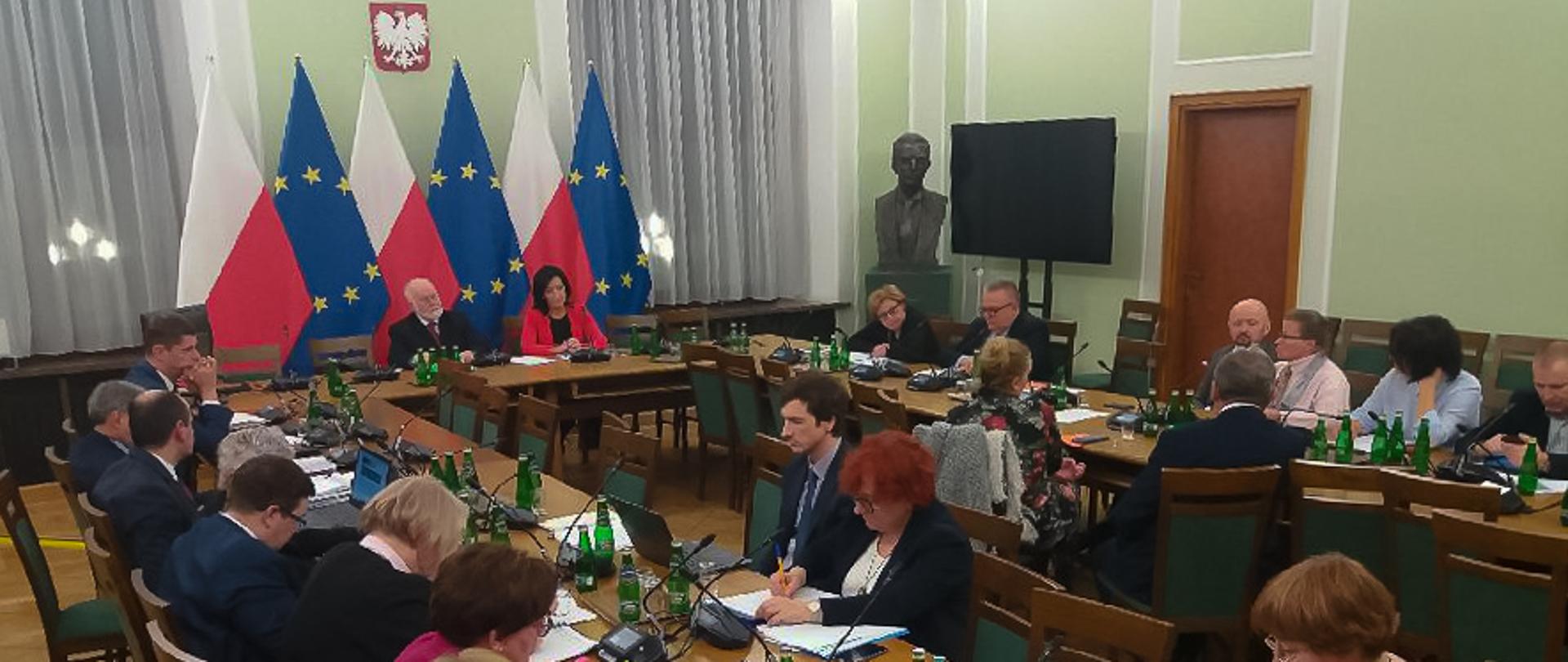 Posiedzenie Komisji Edukacji Nauki i Sportu Senatu RP z udziałem ministra edukacji. Ludzie siedzą przy stołach, pochylają się nad dokumentami i dyskutują. W tle flagi Polski oraz Unii Europejskiej.