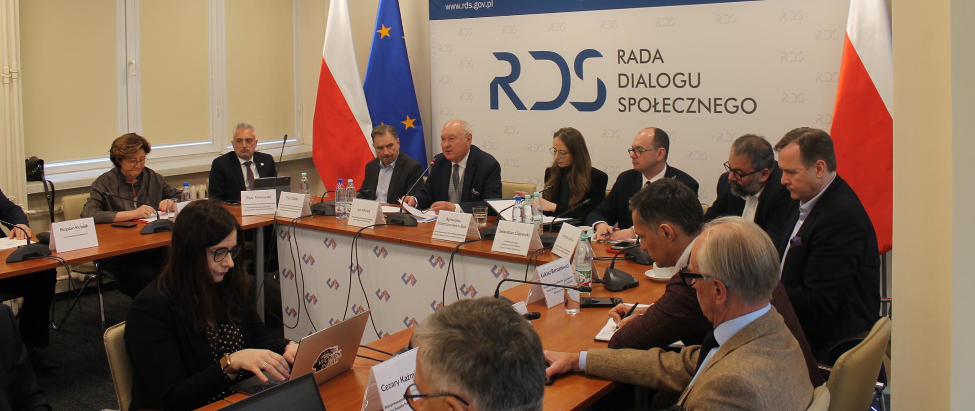 Na zdjęciu widnieje stół prezydialny za którym siedzą od lewej strony: Dorota Gardias – Przewodnicząca Forum Związków Zawodowych, Piotr Ostrowski – Przewodniczący Ogólnopolskiego Porozumienia Związków Zawodowych, Piotr Duda – przewodniczący Rady Dialogu Społecznego, Jan Klimek – Przewodniczący Rady Dialogu Społecznego, Agnieszka Dziemianowicz-Bąk Ministra Rodziny, Pracy i Polityki Społecznej, Sebastian Gajewski – Podsekretarz Stanu w Ministerstwie Ministra Rodziny, Pracy i Polityki Społecznej, Liwiusz Laska – Dyrektor Generalny w MRPiPS oraz pozostali partnerzy. W tle siedzących dwie flagi: UE i Polski oraz baner z napisem Rada Dialogu Społecznego.