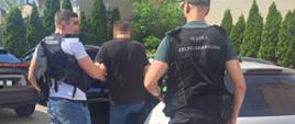 Funkcjonariusze Służby Celno‑Skarbowej prowadzą osobę w kierunku otwartego samochodu.