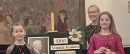Na zdjęciu kobieta blondynka w okularach i czarnej sukience. Przed nią dwie uczennice. Obie trzymają skrzypce ze smyczkami. Uczennica po lewej stronie blondynka w czarnej bluzce i beżowych spodniach. Po prawej stronie zdjęcia dziewczynka w różowej sukience. Między uczennicami stojak z napisem "XXVI Sopocki Kon kurs Skrzypcowy im. Jerzego Hazuki" i zdjęciem Jerzego Hazuki. Na ziemi ciemny dywan i brązowa makieta na ścianie.