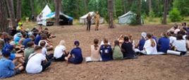 Zdjęcie przedstawia obozowiczów zgromadzonych podczas przeprowadzonej pogadanki w tle widać namioty harcerskie oraz las.