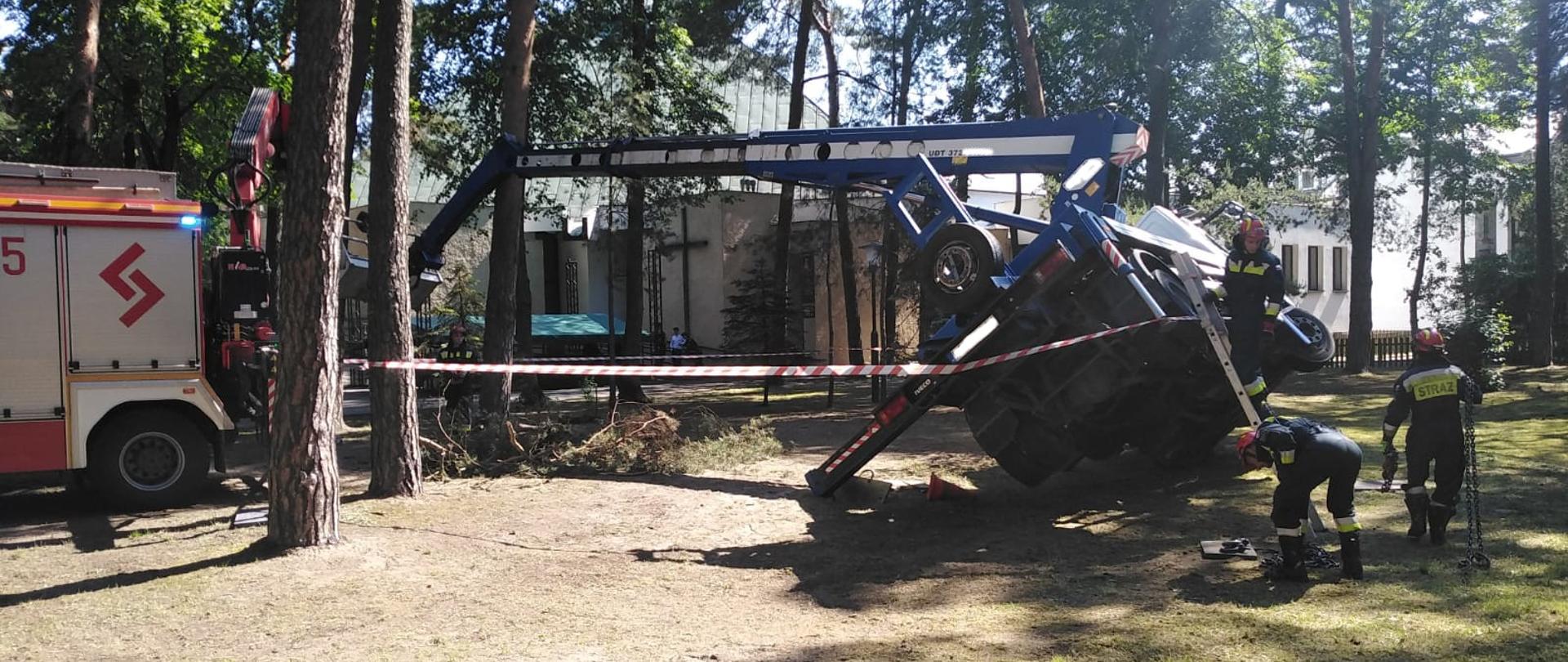 Na zdjęciu pochylony podnośnik koszowy oraz pojazd specjalistyczny państwowej straży pożarnej podczas tworzenia układu do postawienia podnośnika. W tle dwaj strażacy z grupy specjalistycznej ratownictwa technicznego.