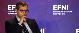 Wiceminister rozwoju i technologii Marek Niedużak siedzący w fotelu podczas obrad EFNI'21 w Sopocie, za jego plecami baner w kolorach czerwieni i niebieskiego z logo EFNI