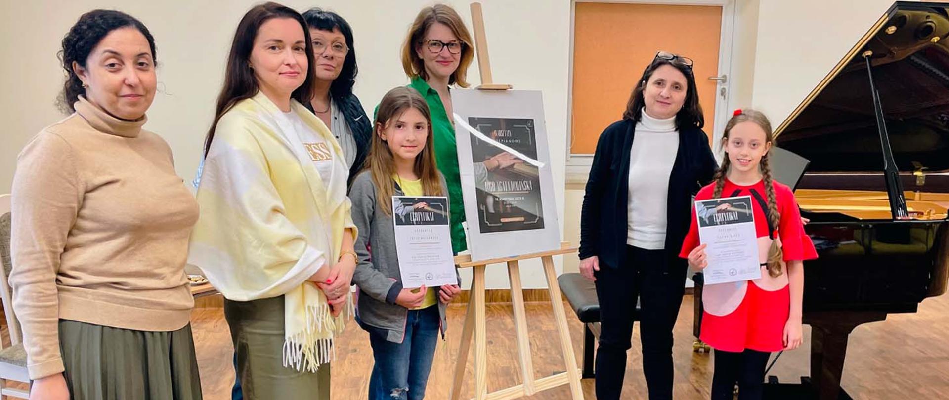 Uczestnicy warsztatów pozują do zdjęcia, pięć kobiet, dwie dziewczynki, pośrodku nich stoi sztaluga z plakatem reklamującym warsztaty, po prawej stronie widoczny fortepian.