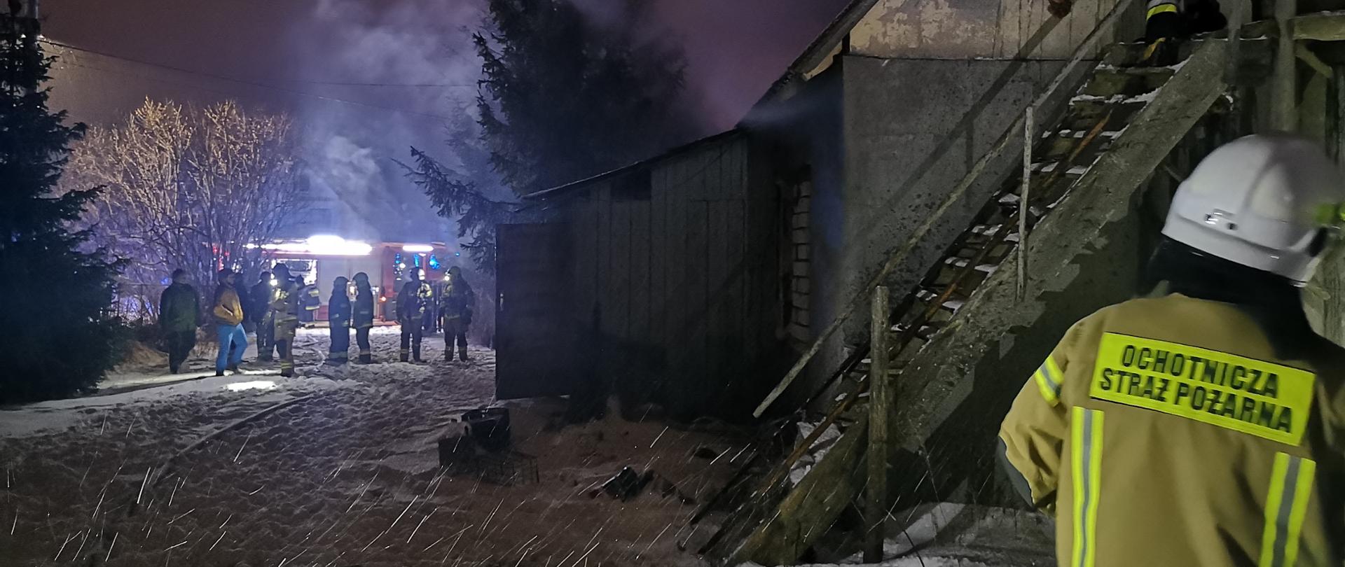 Zdjęcie przedstawia budynek w którym doszło do pożaru. Nad budynkiem unosi się biały dym. W oddali widać samochody straży pożarnej oraz strażaków. Na schodach prowadzących na strych budynku znajduje się strażak sprawdzający miejsce po pożarze. 