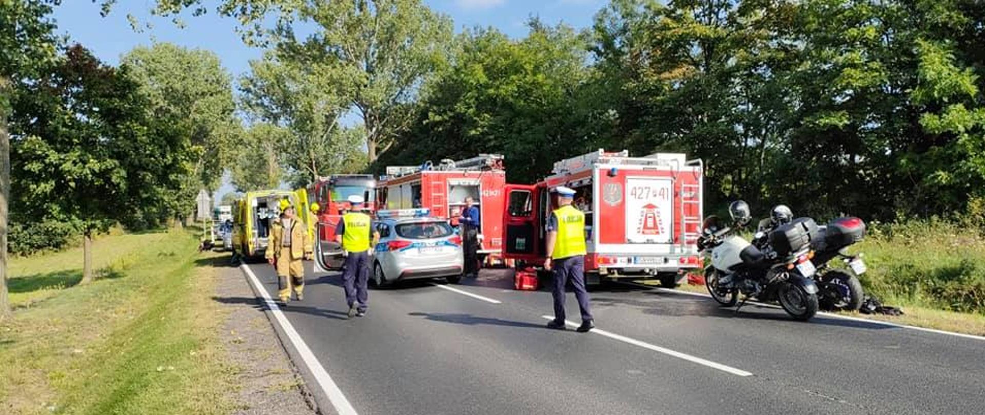 Słonecznie. Asfaltowa czarna droga, na niej czerwone samochody straży pożarnej, motocykle Policji, żółta karetka. Policjanci w granatowych mundurach prowadzą czynności.